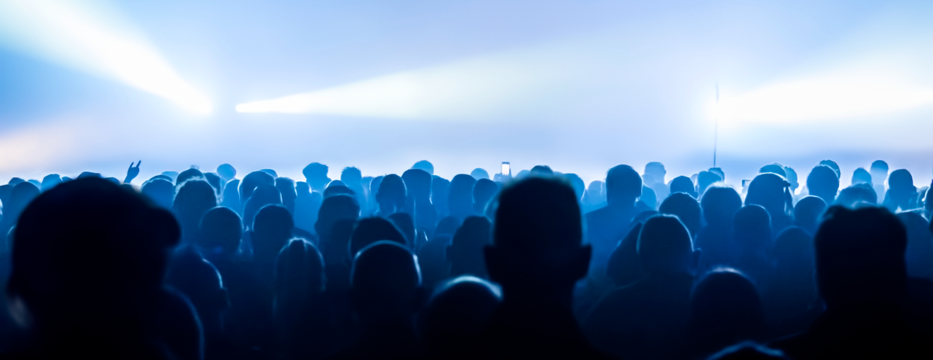 Crowd of people silhouetted in blue lighting at a concert or live event with bright stage lights in the background