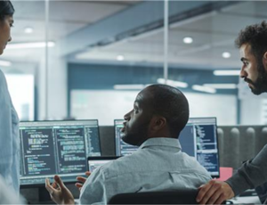 Team of IT professionals collaborating at multiple computer screens displaying code in a modern office