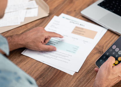 Person reviewing bills and using a calculator app on a smartphone while sitting at a table with documents and a laptop