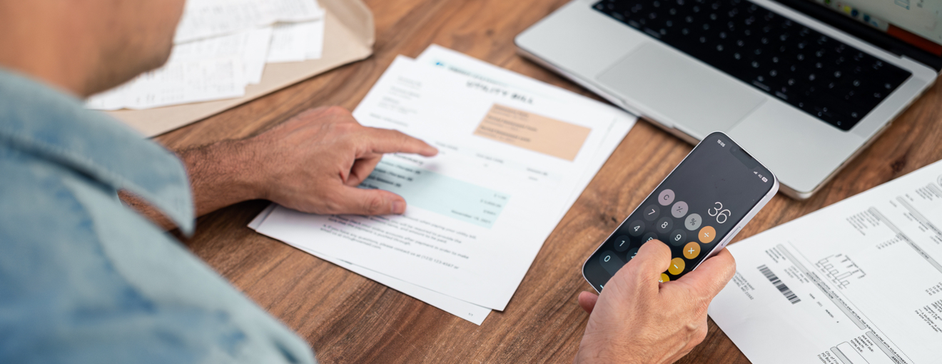 Person reviewing bills and using a calculator app on a smartphone while sitting at a table with documents and a laptop