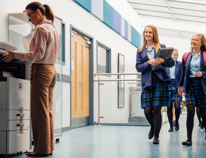 Students in school uniforms walking down a hallway while a staff member uses a printer