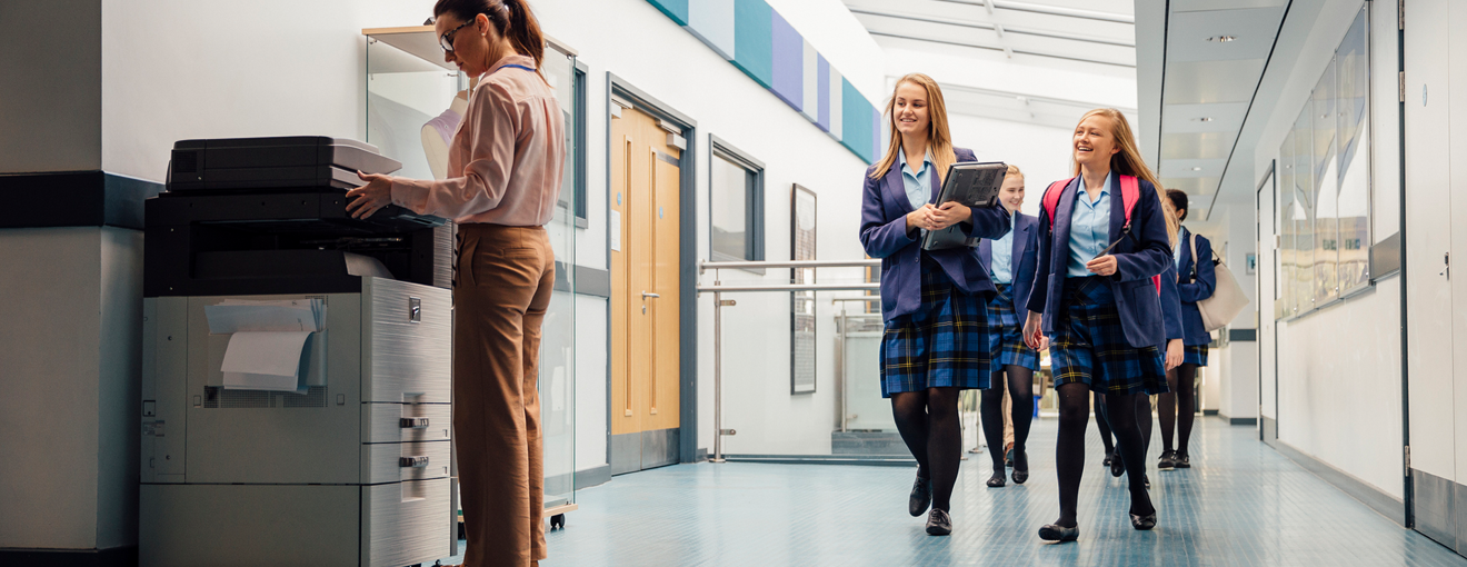 Students in school uniforms walking down a hallway while a staff member uses a printer