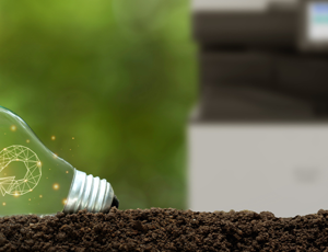 Light bulb lying on soil with a circular sustainability symbol glowing inside, with a blurred office printer in the background